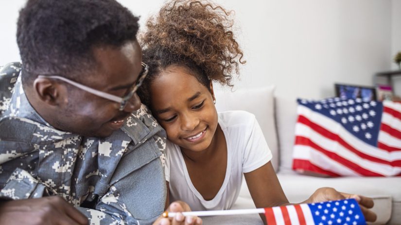 Portrait of happy american family father in military uniform and cute little girl daughter with flag of United States hugging and smiling at camera, male soldier dad reunited with family at home