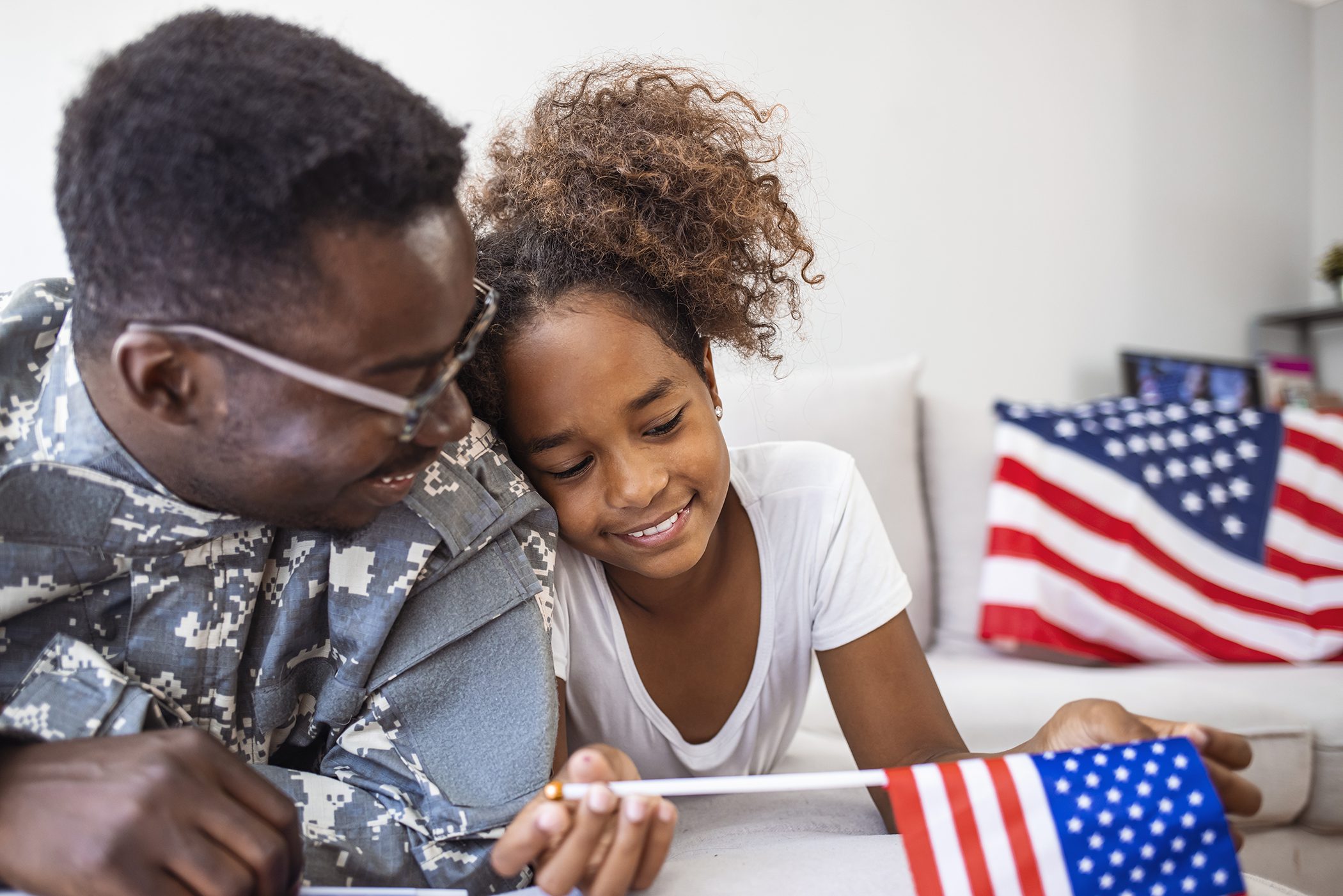 Portrait of happy american family father in military uniform and cute little girl daughter with flag of United States hugging and smiling at camera, male soldier dad reunited with family at home