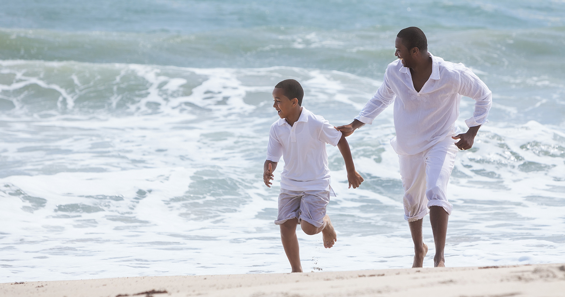 A happy African American family of father and son, man & boy child, running and having fun in the sand and waves of a sunny beach