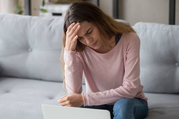 Stressed exhausted upset millennial woman put head on hand, suffering from sudden strong throbbing head ache due to computer remote overworking, feeling tired, sitting on sofa in living room at home.