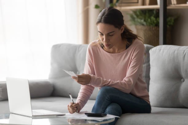 Serious young woman calculating counting vat paying domestic bills writing notes at home, focused female renter do paperwork planning expenses using calculator laptop managing finances sit on sofa