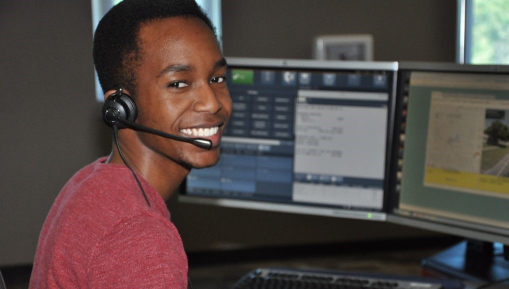 A Pinellas County 9 1 1 telecommunicator wearing his headset and sitting in front of his computer screens smiles at the camera.