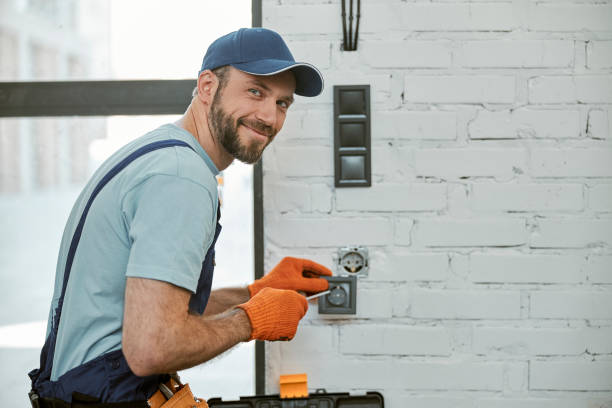 Bearded male electrician looking at camera and smiling while repairing power electric socket with screwdriver