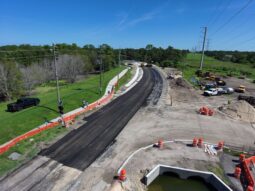 A picture of the newly paved road approaching the new bridge on Old Coachman Road. The road curves to the left to the new bridge, and there are piles of dirt, construction barriers and construction equipment.
