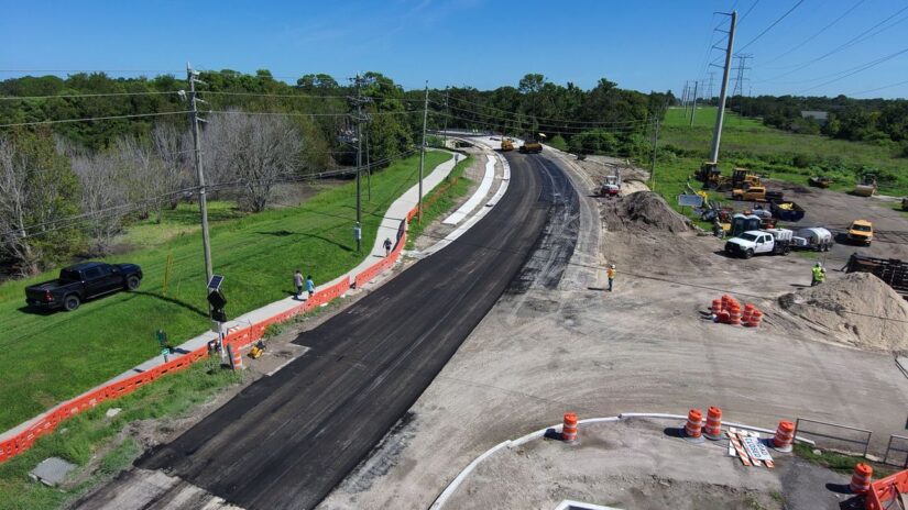 A picture of the newly paved road approaching the new bridge on Old Coachman Road. The road curves to the left to the new bridge, and there are piles of dirt, construction barriers and construction equipment.