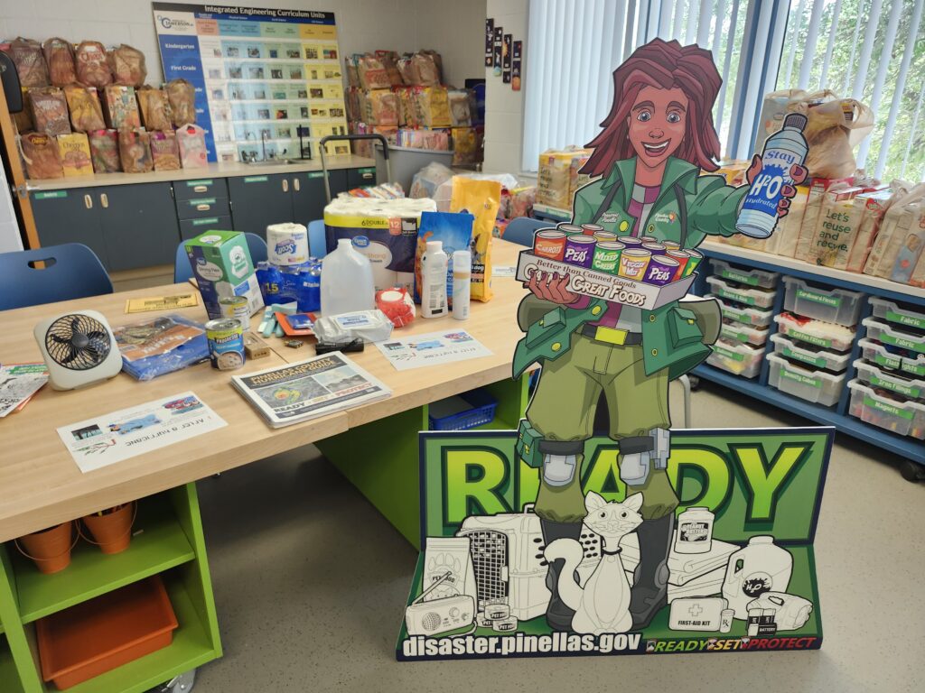 A cut-out character holding hurricane supplies is in front of supplies in a classroom.