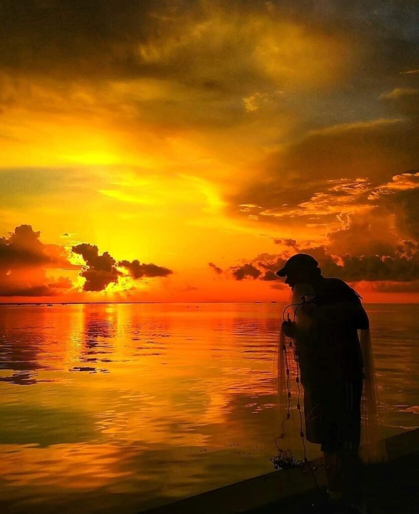 A photo of a silhouette of a man standing on the short of a bay holding a fishing net in the early morning