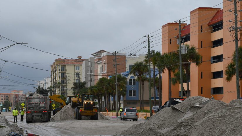 A Johns Pass area street covered with piles of sand while utility workers and vehicles remove sand