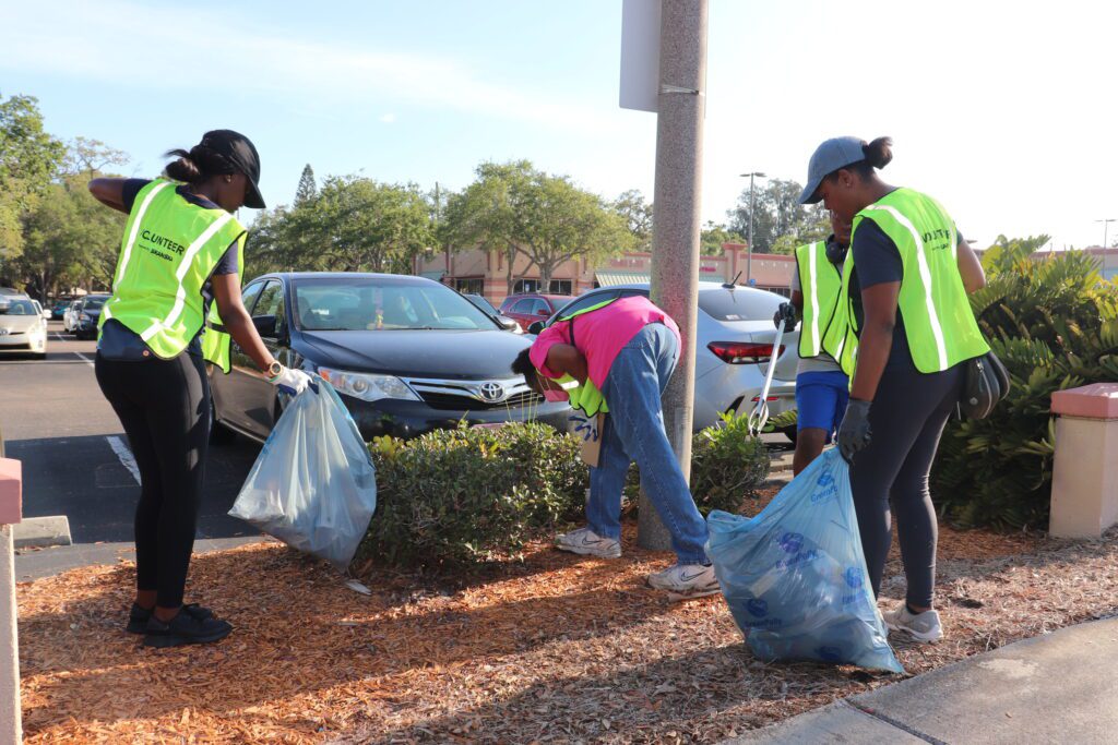 A small group of people using pickers to collect litter in the mulch next to the parking lot.