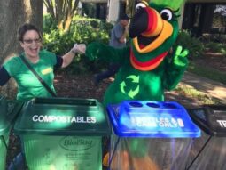 Person and someone in a bird costume standing behind a recycling and compost bin.