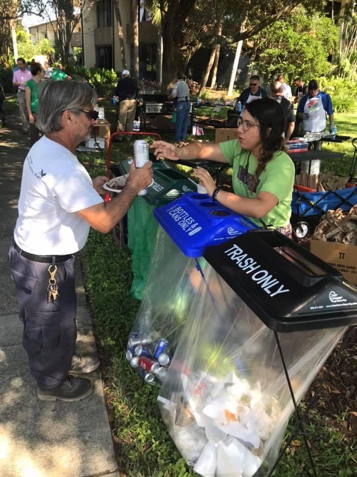 A person handing an empty metal over three waste cans to another person who is helping separate waste into recycling, compost and trash.