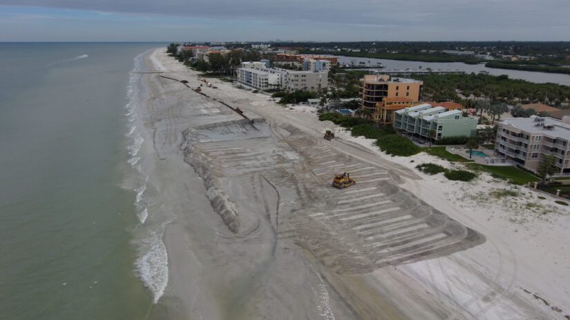 Arial photo of Indian Shores beach with water on the left and buildings on the right. A pipeline is shown with ongoing beach construction of widening the beach.