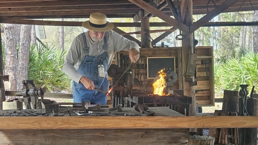 Jay Dugan, an artisan blacksmith, works at an outdoor forge surrounded by rustic wooden beams and brick walls. He wears a straw hat, gray shirt, and denim overalls, shaping heated metal with a hammer as flames rise from the forge.