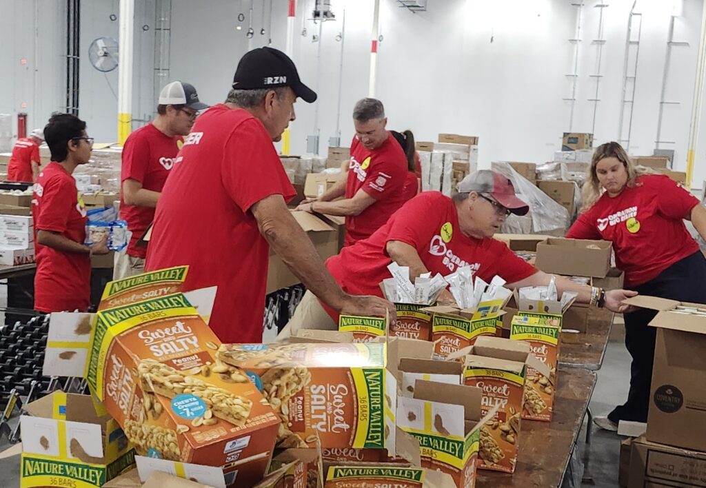 Several workers are in two rows packing boxes with granola bars and other food supplies.