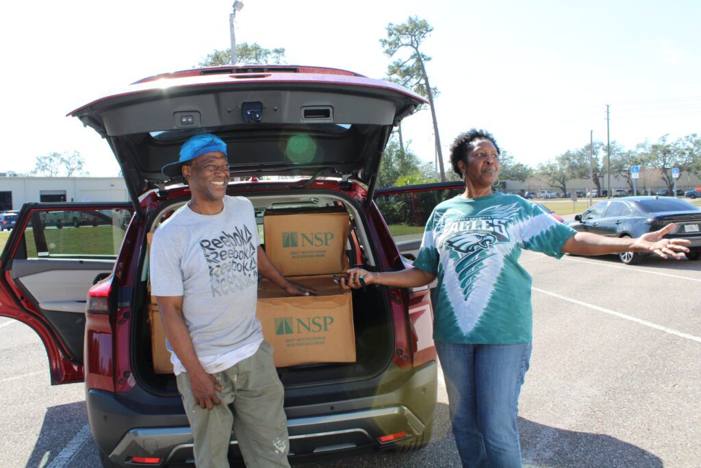 A smiling man and woman stand at the back of their car with the truck open, showing several boxes.
