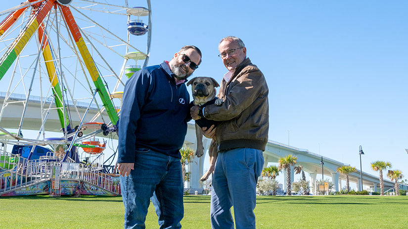 Chairman Eggers and Commissioner Latvala hold K-9 Gucci in front of a ferris wheel in Coachman Park.