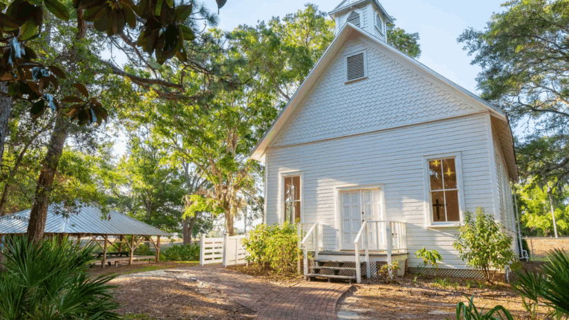 A front-angle view of the historic Safety Harbor Church, a white frame vernacular building preserved at Heritage Village in Largo, Florida. The church features a steep gabled roof with decorative imbricated (scalloped) shingles on the pediment, a small belfry, and double wooden front doors. A curved brick path leads to the entrance, flanked by lush oak trees, palms, and a nearby open-air wooden pavilion.