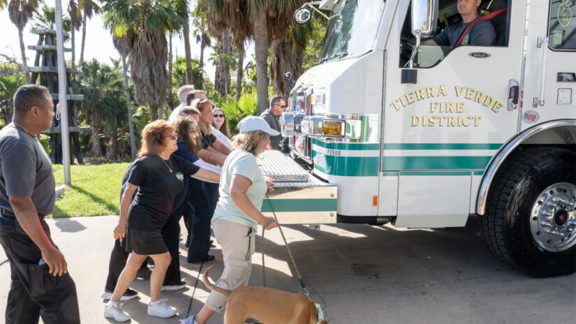 A group of community members push a new Tierra Verde Fire District fire engine into a fire station bay during a ceremonial “truck push.” A firefighter sits in the driver’s seat while several people push from the side of the white and green engine.
