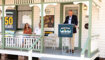 Commissioner Brian Scott speaks at a Heritage Village podium on the porch of the historic white-clapboard Plant-Sumner House during the "From Dedication to Designation" ceremony at Heritage Village in Largo on March 28, 2026. Seated behind him are Monica Drake and Commissioner Chris Latvala. To the left, a large vertical banner celebrates Heritage Village's 50th anniversary (1976–2026) alongside a historical display board.