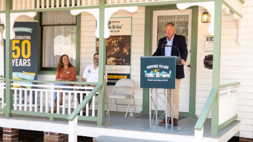 Commissioner Brian Scott speaks at a Heritage Village podium on the porch of the historic white-clapboard Plant-Sumner House during the "From Dedication to Designation" ceremony at Heritage Village in Largo on March 28, 2026. Seated behind him are Monica Drake and Commissioner Chris Latvala. To the left, a large vertical banner celebrates Heritage Village's 50th anniversary (1976–2026) alongside a historical display board.