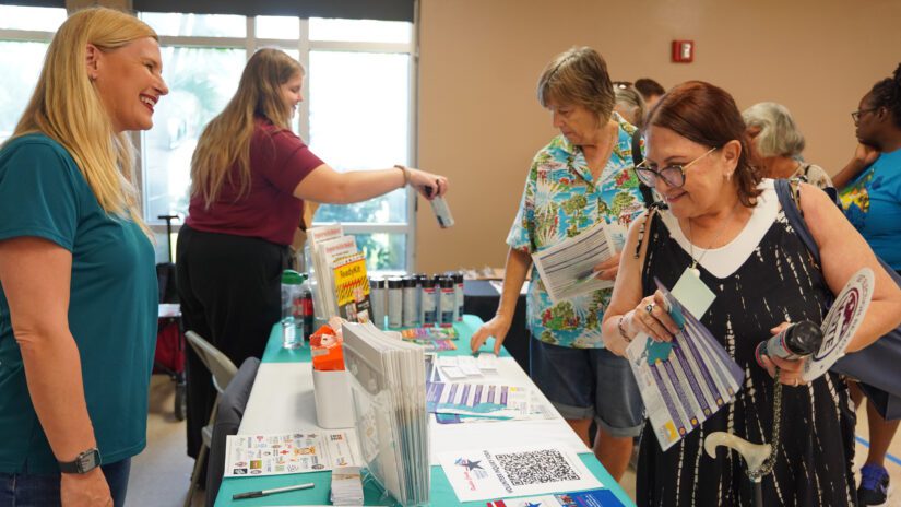 Attendees at Pinellas County’s Hurricane Preparedness Day expo gather around an information table where staff share emergency preparedness materials, brochures, and supplies. A staff member smiles while speaking with visitors as others review printed guides and pick up preparedness items.