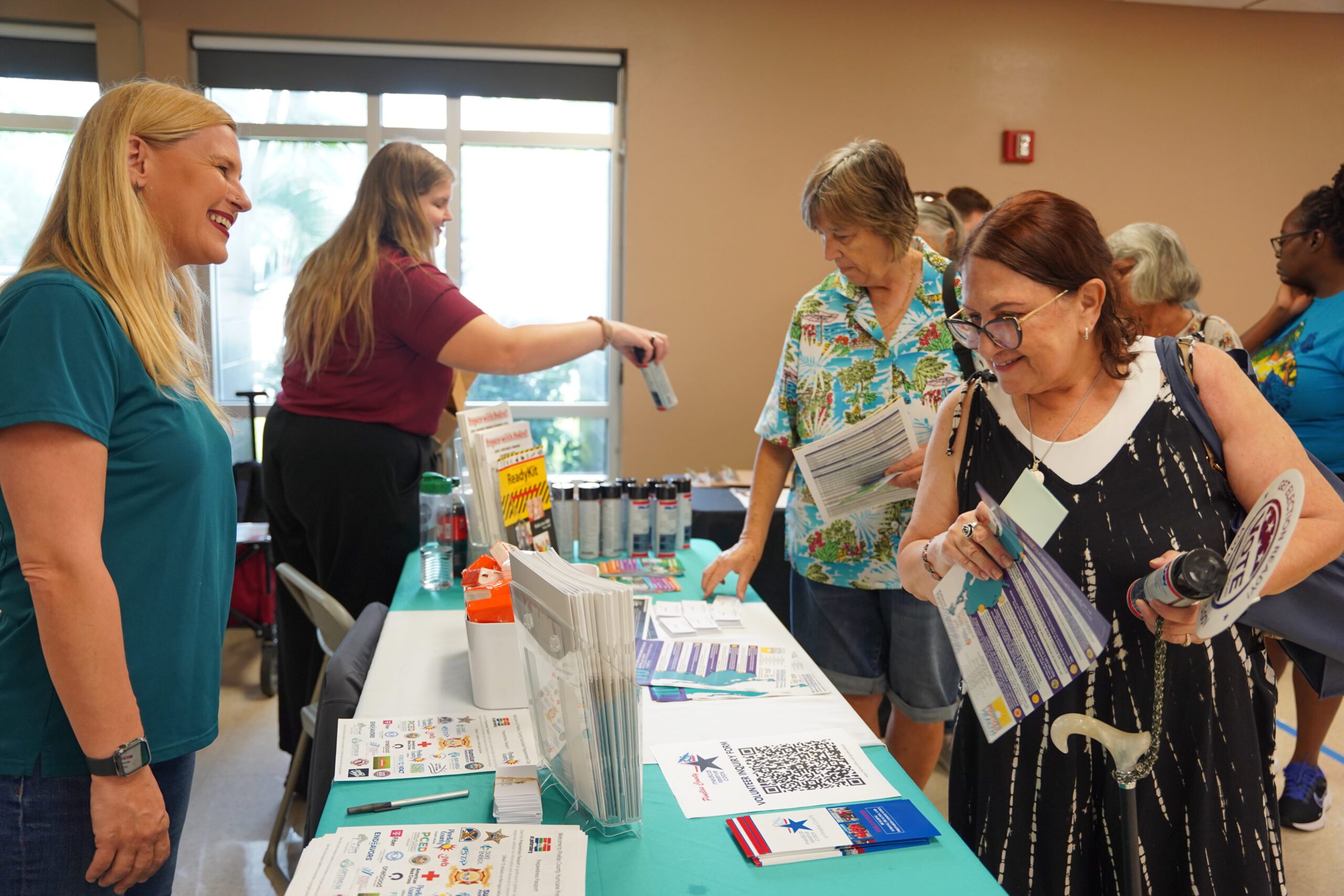 Attendees at Pinellas County’s Hurricane Preparedness Day expo gather around an information table where staff share emergency preparedness materials, brochures, and supplies. A staff member smiles while speaking with visitors as others review printed guides and pick up preparedness items.