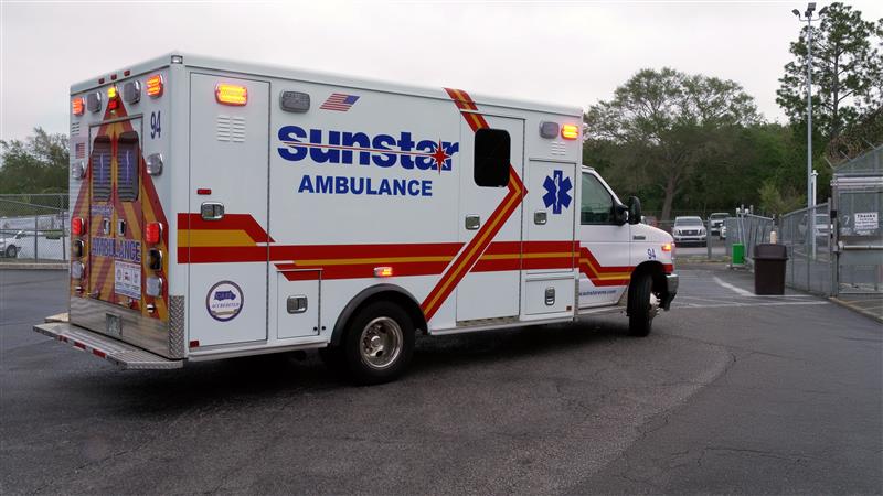 A Sunstar ambulance is shown with lights on driving from left to right towards a chain linked gate. Cars are seen in a parking lot in the background.