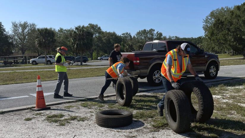 Staff wearing reflective safety vests collect and move discarded tires at a Pinellas County free tire disposal event. Two people roll large tires away from a pickup truck, while another looks at a clipboard. A traffic cone and parked vehicles are visible nearby on a sunny day.
