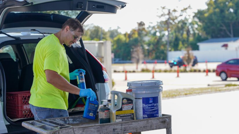 A worker wearing gloves and a bright safety shirt unloads household hazardous waste items from the back of a vehicle during a Pinellas County Household Hazardous Waste North chemical collection event. Several paint cans and chemical containers are placed on a rolling cart in the foreground, with traffic cones, trees, and parked vehicles visible in the background.