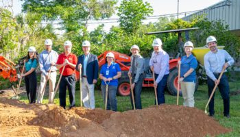 People posing with shovels and hardhats with an excavator in the background.