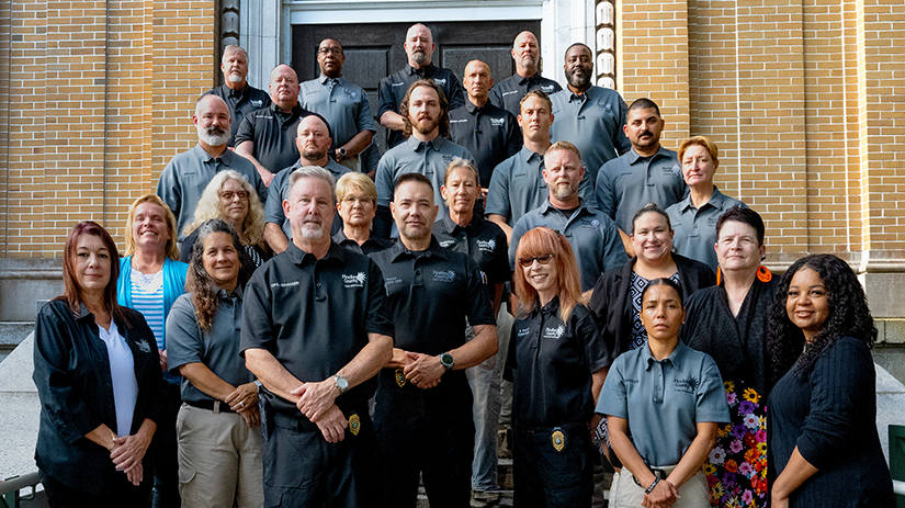 A group photo of approximately 24 members of the Pinellas County Code Enforcement Team for the file Code Enforcement Week_5-19-2026_16-9_AR.jpg. A diverse group of men and women are arranged in staggered rows on the concrete steps leading up to the dark double doors of a tan brick building. Most team members are wearing official gray or black polo shirts featuring the Pinellas County logo, with some displaying enforcement badges. They are facing forward, looking at the camera.