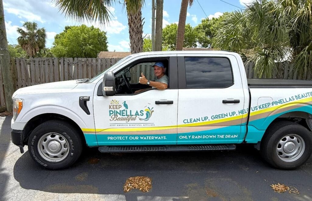 A white pickup truck with “Keep Pinellas Beautiful” branding is parked in a paved lot lined with palm trees. A staff member sits in the driver’s seat, smiling and giving a thumbs-up through the open window. Environmental messages promoting clean waterways and community beautification are displayed along the side of the truck.