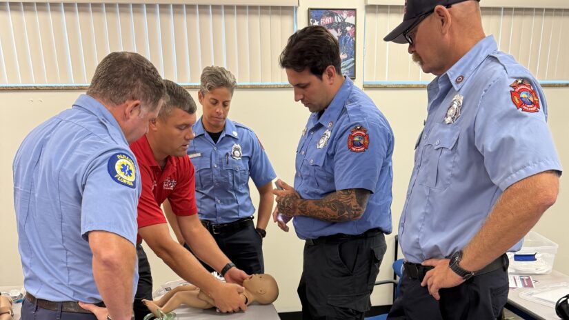 Five emergency medical personnel stand around a table during a pediatric emergency care training session. An instructor demonstrates techniques on an infant medical mannequin while the group observes closely.