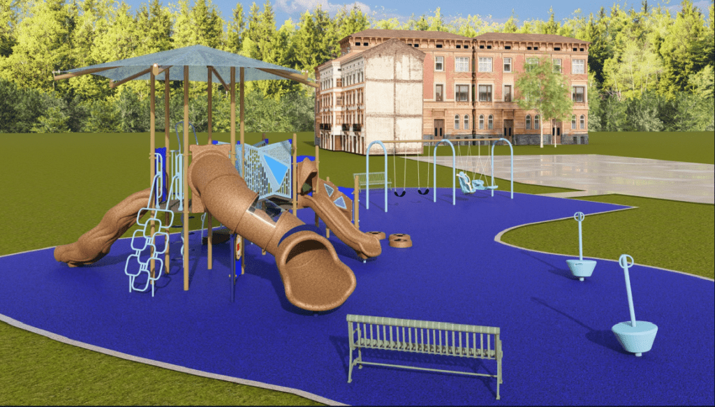 The image shows an outdoor playground built on a bright blue rubberized safety surface with a curved, irregular outline. The playground is located in a large grassy area with a dense forest of tall green trees in the background. Several multi‑story brick buildings are visible beyond the treeline.
In the foreground, a green metal bench faces the play area. The central feature of the playground is a large multi‑level play structure. The structure includes several elevated platforms connected by ramps, stairs with handrails, and climbing components such as a geometric metal climbing ladder. A large brown enclosed tube slide extends from an upper platform and curves downward. A second open slide, also brown, is positioned to the right. A square roof canopy supported by tall posts provides shade over the central portion of the structure.
To the right of the main structure is a swing set with a light‑blue metal frame. The swing set includes two belt swings and two molded adaptive swings with supportive seats. Several stepping pods, arranged in a small group, are placed near the base of the slides.
On the far right, two small cup‑shaped spinning play elements sit on individual poles on the blue safety surface.
The sky is bright with scattered clouds, indicating sunny weather and clear visibility.