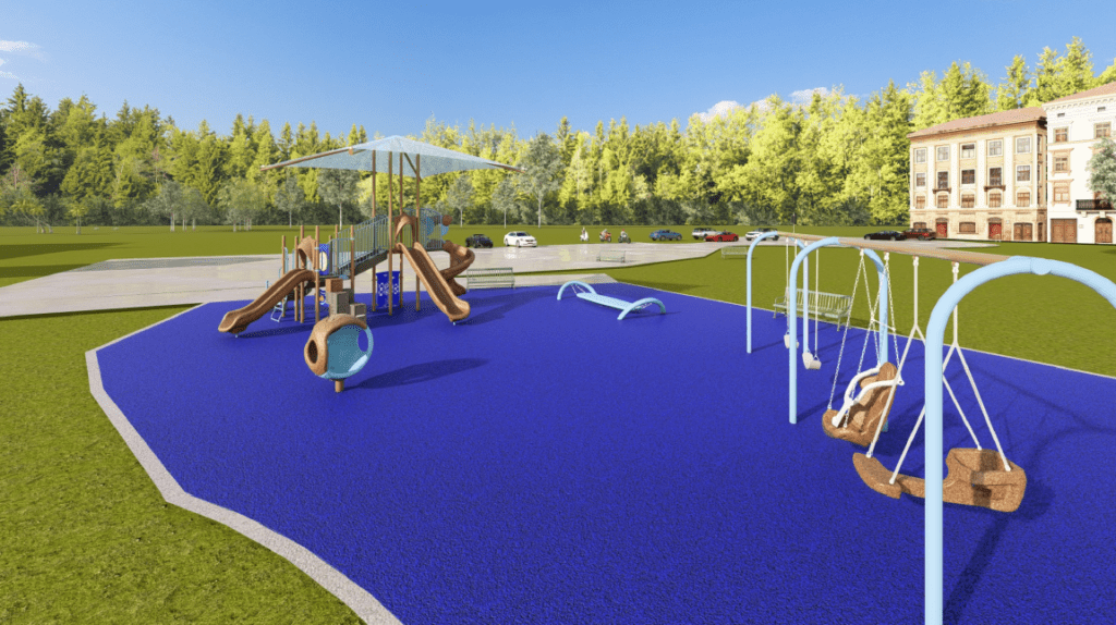 The image shows an outdoor playground situated on a bright blue, rubberized safety surface with an irregular curved boundary. In the center-left of the playground is a large multi‑level play structure with several ramps, handrails, activity panels, and multiple slides. The slides are brown and include both straight and curved options. Above the main structure is a wide square shade canopy supported by tall posts. A rounded, spherical climbing or activity element with openings is positioned near the front of the structure.
On the right side of the playground is a swing set with several swings, including two bucket‑style swings and one standard belt swing. A long, low balancing or rocking structure painted light blue sits near the center-right area of the play surface. A green bench is located behind the swings.
In the background, there is a paved parking area with several parked vehicles. Beyond the parking area is a dense treeline of tall green trees extending across the horizon. On the far right, a multi‑story light‑colored brick building is partially visible. The sky is bright blue with a few small clouds, indicating clear weather.