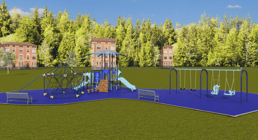 The image shows an outdoor playground installed on a bright blue rubberized safety surface with a curved, irregular outline. The playground is set within a large grassy field bordered by tall, dense trees in the background. Several multi‑story brick buildings are spaced across the horizon behind the treeline.
The playground contains multiple play features. On the left side, there is a large rope‑climbing structure shaped like a wide pyramid made of interconnected ropes supported by metal poles. Adjacent to the rope structure, near the center, is a multi‑level play structure with platforms, ramps, railings, and activity panels. This structure includes a covered roof canopy, a small climbing wall, and two light‑blue slides extending toward the right. Near the front of the play structure, there are three small spring rider toys shaped like abstract animals or figures.
On the far right side of the playground is a swing set with several swings: two bucket‑style swings, one standard belt swing, and one adaptive swing with a high‑back molded seat. Two green metal benches face the play area—one positioned on the left side near the rope‑climbing structure and another near the center‑right beside the slides.
The sky is clear and blue, and the lighting suggests bright, sunny weather.