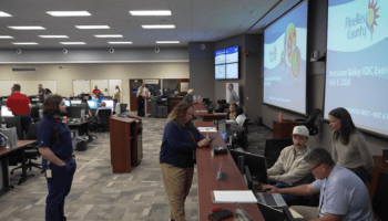 Wide view of Pinellas County's Emergency Operations Center during the countywide Hurricane Bailey exercise. Staff from multiple departments work at computer stations and collaborate in small groups. Large screens share information about the exercise in the background.
