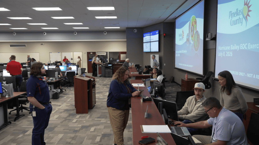 Wide view of Pinellas County's Emergency Operations Center during the countywide Hurricane Bailey exercise. Staff from multiple departments work at computer stations and collaborate in small groups. Large screens share information about the exercise in the background.