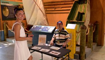 A smiling woman in a white sleeveless dress stands in front of a black folding table, looking toward the camera. Across from her, a man wearing a striped polo shirt and a blue baseball cap is seated in a folding chair behind the table. They are inside the McKay Creek Boat Shop. Behind them, historical maritime exhibits are on display, including a small rigged sailboat on the left and a weathered, green-and-white rowboat propped up vertically behind wooden pilings on the right.