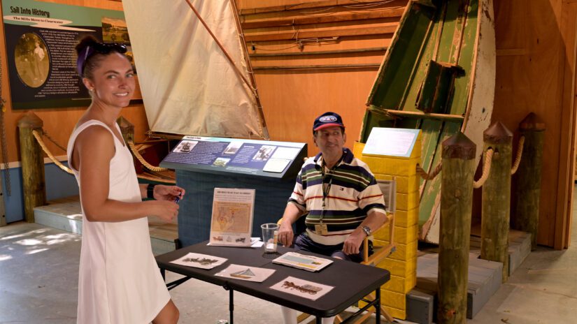 A smiling woman in a white sleeveless dress stands in front of a black folding table, looking toward the camera. Across from her, a man wearing a striped polo shirt and a blue baseball cap is seated in a folding chair behind the table. They are inside the McKay Creek Boat Shop. Behind them, historical maritime exhibits are on display, including a small rigged sailboat on the left and a weathered, green-and-white rowboat propped up vertically behind wooden pilings on the right.