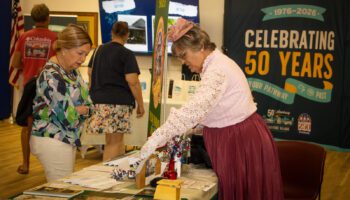 A woman dressed in vintage attire with a lace blouse and pink fascinator hat presents historical display items to a visitor at an informational table.