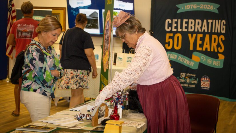 A woman dressed in vintage attire with a lace blouse and pink fascinator hat presents historical display items to a visitor at an informational table.