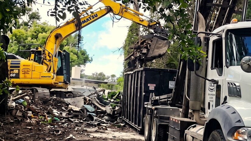 A yellow Komatsu excavator uses its arm to load structural debris into a large black dumpster attached to a white commercial truck. The outdoor demolition site is surrounded by trees and foliage, with piles of rubble scattered across the dirt ground.
